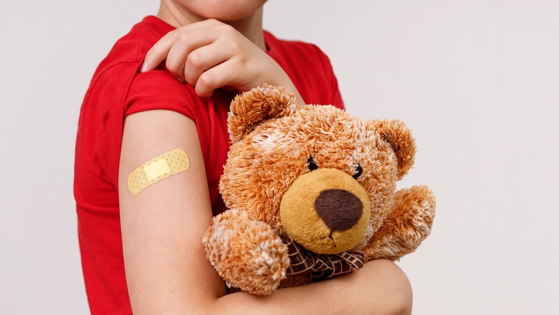 Children in red t-shirt holds toy and shows his shoulder after vaccination. Child holding a bear showing off the bandage on his arm after he got is vaccine shot.